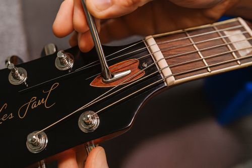 Close-up of a person adjusting the truss rod on a Les Paul–style guitar headstock using a metal wrench, showing the glossy black headstock, tuning pegs, and truss rod cover removed.