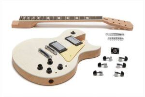 Unfinished Les Paul–style DIY guitar kit laid out on a white background, including the carved-top body, separate neck with inlays, humbucker pickups, bridge, tuners, and hardware components.